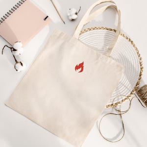 Puede incluir: Bolso tote de color crema con un logotipo de llama roja. El bolso tiene dos asas y est&aacute; hecho de una tela resistente. El bolso est&aacute; sobre una superficie blanca con un cuaderno, un l&aacute;piz, ramitas de algod&oacute;n y un salvamanteles tejido.