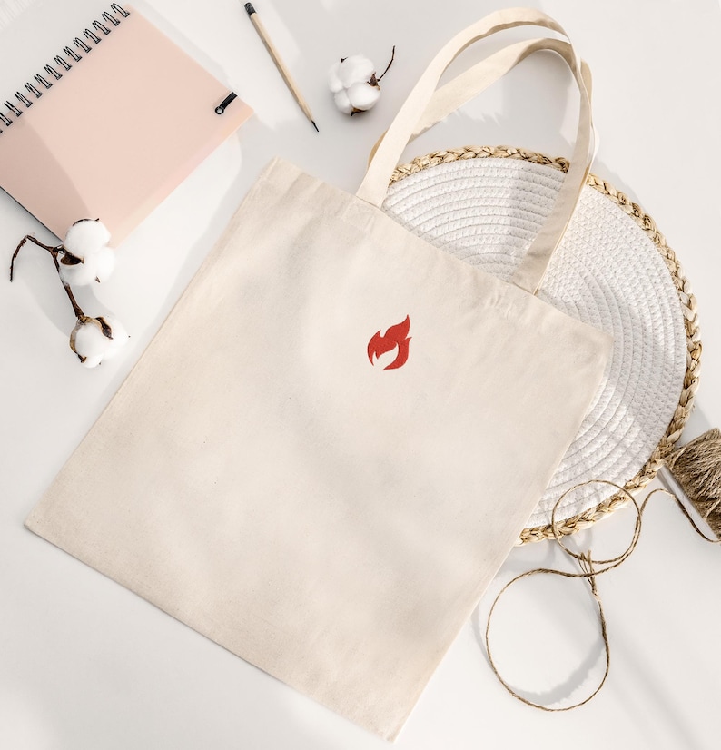 Puede incluir: Bolso tote de color crema con un logotipo de llama roja. El bolso tiene dos asas y est&aacute; hecho de una tela resistente. El bolso est&aacute; sobre una superficie blanca con un cuaderno, un l&aacute;piz, ramitas de algod&oacute;n y un salvamanteles tejido.