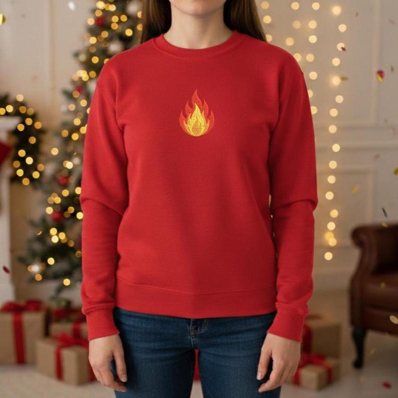 Puede incluir: Una sudadera roja con una llama bordada en el pecho. El dise&ntilde;o de la llama presenta colores amarillos y naranjas. La sudadera es de color liso y tiene mangas largas. El fondo incluye un &aacute;rbol de Navidad y regalos envueltos.