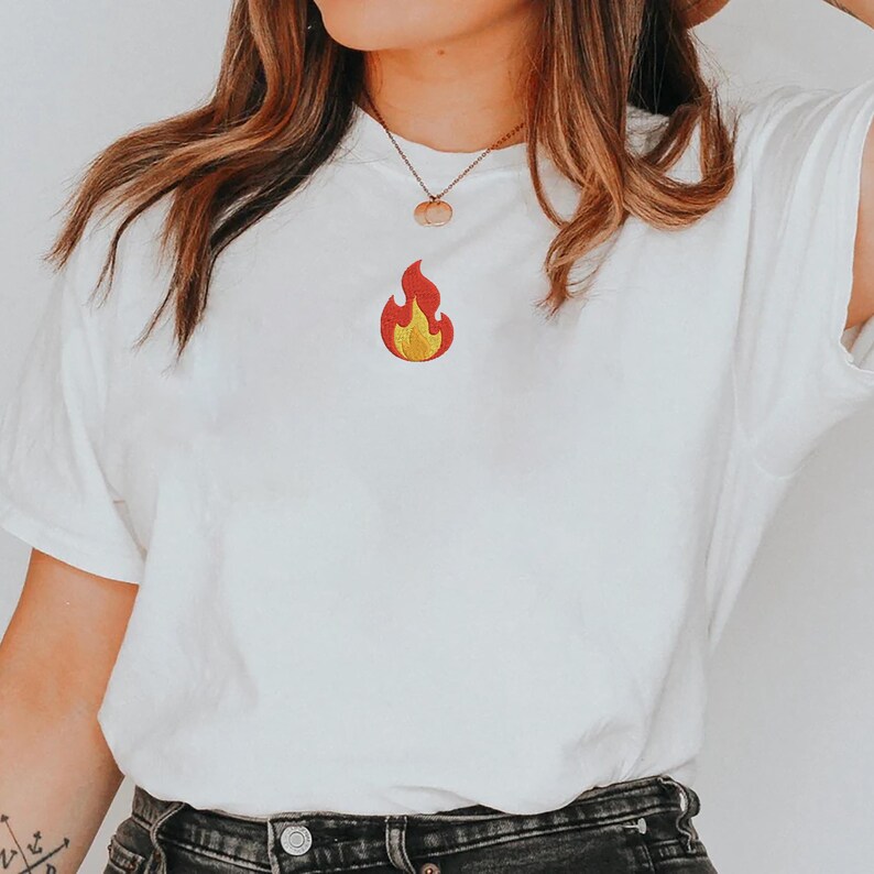 Puede incluir: Una camiseta blanca con un peque&ntilde;o dise&ntilde;o de llama bordada en rojo y amarillo. La llama est&aacute; centrada en el pecho. La camiseta est&aacute; hecha de un material suave y tiene mangas cortas.