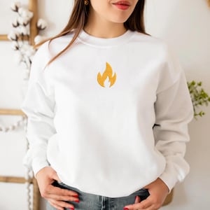 Puede incluir: Una sudadera blanca con un peque&ntilde;o dise&ntilde;o de llama amarilla bordada centrada en el pecho. La parte superior de manga larga y cuello redondo est&aacute; hecha de un material suave y es adecuada para ropa casual. La sudadera tiene un dise&ntilde;o simple y minimalista.