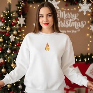 Puede incluir: Una sudadera blanca con un peque&ntilde;o dise&ntilde;o de llama bordada. La sudadera tiene mangas largas y cuello redondo. El fondo incluye un &aacute;rbol de Navidad y un letrero que dice "Merry Christmas".