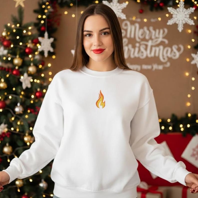 Puede incluir: Una sudadera blanca con un peque&ntilde;o dise&ntilde;o de llama bordada. La sudadera tiene mangas largas y cuello redondo. El fondo incluye un &aacute;rbol de Navidad y un letrero que dice "Merry Christmas".