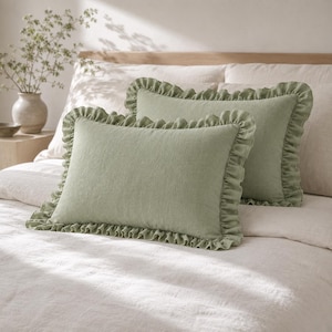 Könnte beinhalten: Zwei rechteckige salbeigrüne Dekokissen mit gerüschten Kanten. Die Kissen haben eine strukturierte Stoffoberfläche und liegen auf einem Bett mit heller Bettwäsche. Das Design umfasst einen gerafften Rüschenrand um den gesamten Umfang.