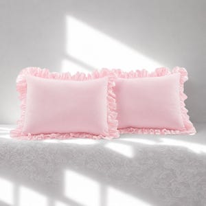 May include: Two rectangular light pink pillows featuring a ruffled border along all four edges. The pillows have a smooth, solid-coloured centre panel and a gathered fabric trim that creates a decorative frill around the entire perimeter.