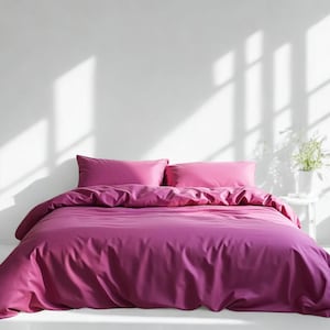 May include: A bed dressed with a magenta duvet cover and two matching pillows. The bedding is arranged on the bed, with shadows cast across the white wall behind it. A small white table with a plant is to the right.