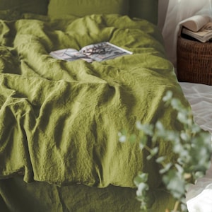 Può includere: Un letto coperto da biancheria da letto in lino verde oliva. La biancheria da letto comprende un copripiumino e federe, con un libro aperto sopra. Un cesto di vimini e lenzuola bianche sono visibili sullo sfondo.