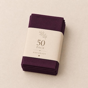 Op de afbeelding: Een stapel van vijftig dieppaarse stoffen servetten, bijeengehouden door een beige papieren band. De band bevat een kleine botanische illustratie, de tekst 50 PACK en het woord HANDMADE met een klein harticoontje eronder.