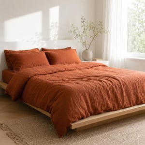 May include: A bed featuring a rust-colored duvet cover and two matching rectangular pillowcases. The bedding has a textured appearance and is placed on a low-profile wooden bed frame. The setup includes a white headboard and a light-colored rug.