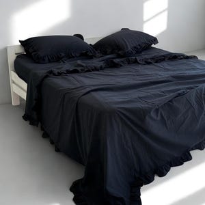 Könnte beinhalten: Ein schwarzes Bettwäsche-Set mit Rüschenkanten. Das Set beinhaltet ein Spannbetttuch, ein Bettlaken und zwei Kissenbezüge. Das Bett steht auf einem weißen Holzrahmen, und die Bettwäsche ist ordentlich angeordnet. Der Raum hat ein minimalistisches Design mit weißen Wänden.