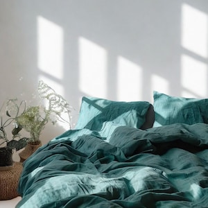 May include: A teal linen bedding set, comprising a duvet cover and two pillowcases, is displayed on a bed. The bedding has a relaxed, slightly creased appearance. The room is lit by natural light, casting shadows on the white wall.