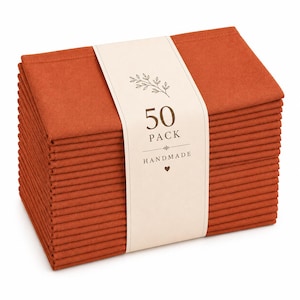 May include: A stack of fifty rectangular burnt orange fabric napkins secured with a cream-colored paper band. The band features a botanical illustration, the text 50 PACK, the word HANDMADE, and a small heart icon printed in dark brown ink.
