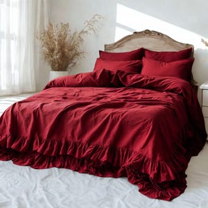 May include: A bed dressed in a deep red duvet cover and pillows. The duvet has a ruffled edge. The headboard is light wood. The bed is in a bright room with white curtains and a white bedside table.