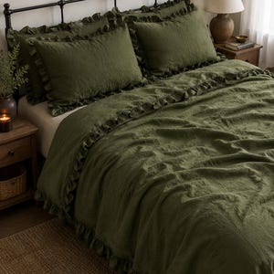 May include: Olive green bedding set with ruffled edges, including a duvet cover and pillow shams. The bed is made up with a dark metal frame, a wooden nightstand, and a lamp. The room has a cozy, inviting atmosphere.