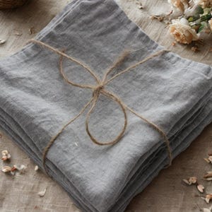Peut inclure: Un ensemble de serviettes en lin gris pliées et attachées avec de la ficelle. Les serviettes sont disposées sur une surface neutre, avec des pétales de fleurs séchées et de petites roses crème éparpillées autour. Les serviettes sont d'un gris doux.