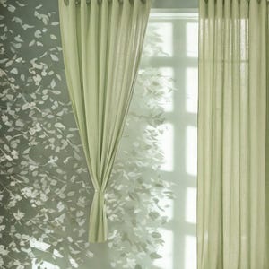 Pode incluir: Cortinas de linho verde claro penduradas em uma barra escura. As cortinas estão parcialmente fechadas, revelando uma janela e uma parede com um padrão de folhas. As cortinas são amarradas com uma fita de tecido combinando.
