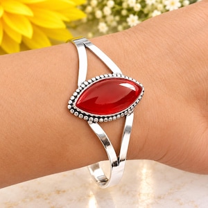 Puede incluir: Una pulsera de plata con una piedra roja central en forma de marquesa. La piedra está rodeada por un borde de metal con cuentas y montada en un diseño de banda dividida. El metal plateado pulido contrasta con la piedra roja lisa y brillante.