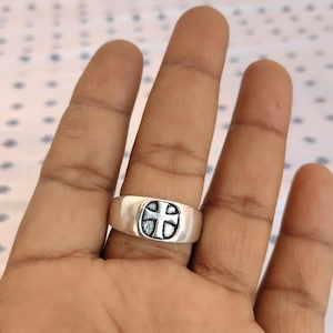 May include: A silver ring featuring a cross motif. The ring has a broad band and an oval face with a detailed cross design. The cross is a lighter colour than the ring's band.