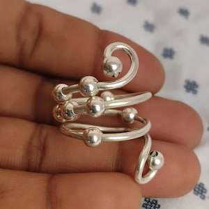 May include: A silver-colored, spiral ring with multiple small, round beads. The ring wraps around the finger in a continuous design, with a smooth, polished finish. The beads are evenly spaced along the spiral, creating a tactile and visually appealing accessory.