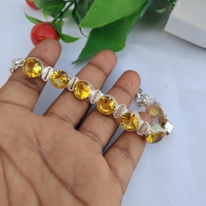 Könnte beinhalten: Ein silbernes Gliederarmband mit einer Reihe runder, facettierter gelber Edelsteine, die in einzelnen Metallfassungen sitzen. Das Armband verfügt über einen Metallverschluss und zeigt ein helles, reflektierendes Finish an den Kettengliedern.