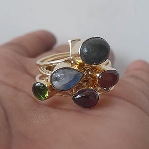 Può includere: Una collezione di anelli d'oro, ognuno ornato da una gemma diversa. Le gemme includono verde scuro, azzurro, rosso-marrone e viola intenso. Gli anelli sono impilati insieme, creando un effetto a strati.