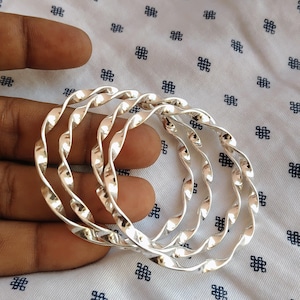 Può includere: Quattro braccialetti color argento con un design a corda intrecciata. I braccialetti sono lisci e lucenti, riflettendo la luce. Lo sfondo è un tessuto bianco con un motivo ripetuto di piccole forme geometriche blu scuro.