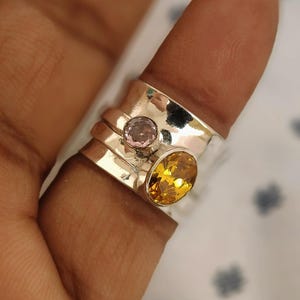 Puede incluir: Un anillo de plata con dos piedras preciosas. Una es una piedra redonda de color rosa claro, y la otra es una piedra ovalada de color amarillo-naranja. El anillo está en un dedo, y el fondo está borroso.