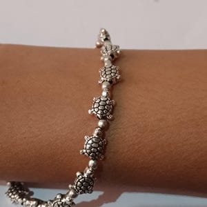 Puede incluir: Una pulsera de tono plateado que presenta un patrón repetitivo de pequeños dijes de tortuga detallados, unidos por diminutas cuentas redondas de plata. El diseño muestra una serie de figuras de tortugas marinas idénticas con caparazones texturizados.