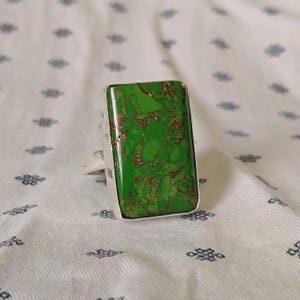 Op de afbeelding: Een rechthoekige ring met een levendige groene steen met een koperkleurig matrixpatroon. De steen is gezet in een gepolijste zilverkleurige metalen band en vertoont een mozaïekachtig uiterlijk met scherpe, hoekige groene en koperen fragmenten.