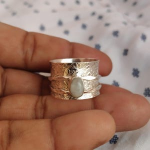 May include: A silver spinner ring featuring an oval, pale stone. The ring has a detailed, textured band with floral designs and two spinning bands. The ring is held in a hand.