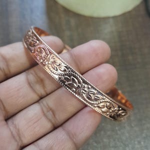 Op de afbeelding: Een koperkleurige armband met een ingewikkeld bloemmotief. De armband is een massieve band met gedetailleerde gravures van bloemen en bladeren. Het patroon herhaalt zich over de gehele omtrek, waardoor een textuur ontstaat.