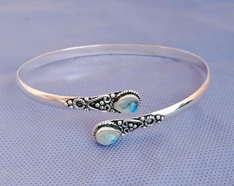 May include: A silver cuff bracelet with detailed patterns and two teardrop-shaped moonstone cabochons. The open-ended bracelet features intricate designs at the ends, with iridescent stones.