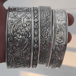Puede incluir: Un conjunto de pulseras plateadas con intrincados diseños florales y de hojas. Las pulseras presentan un diseño en relieve detallado, mostrando varios motivos botánicos. El metal tiene un acabado pulido, reflejando la luz y resaltando los detalles en relieve.