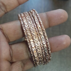 Op de afbeelding: Een set van zes roségouden armbanden met ingewikkelde, zich herhalende patronen van harten, diamanten en bloemmotieven. De armbanden zijn op elkaar gestapeld, wat het gedetailleerde vakmanschap en de warme metallic kleur laat zien.