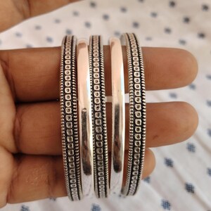 May include: Five silver bangles are shown. Two bangles have a detailed pattern of small circles and lines along the edges. The other three bangles are smooth and polished. The bangles are displayed on a hand.