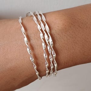 May include: A set of four silver-colored bangles, each featuring a continuous twisted or spiral design. The bracelets are thin, metallic, and have a reflective surface, worn together to create a layered appearance on the wrist.