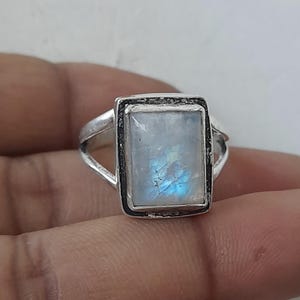 May include: A silver ring featuring a rectangular, iridescent moonstone. The stone displays a play of blue and white colors. The ring has a split shank design.