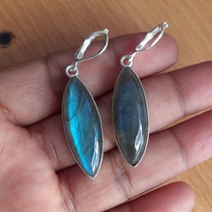Puede incluir: Un par de pendientes plateados con piedras de labradorita ovaladas y alargadas. Las piedras muestran tonos iridiscentes azules y turquesas, engastadas en un marco plateado pulido. Cierre de palanca.