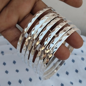 May include: A collection of silver bangles with a hammered finish. The bracelets are circular and polished, reflecting light. The bangles are held in a hand against a white background with a blue pattern.