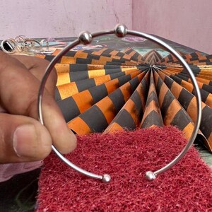 May include: A silver-colored bangle bracelet with small ball accents. The bracelet is held over a red textured surface and a background with black and orange checkered pattern.