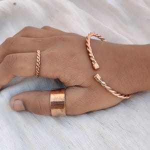 Peut inclure: Une main présente des bijoux en cuivre, dont un bracelet torsadé en forme de corde, une bague assortie et une large bague. Les bijoux ont une teinte brun-rougeâtre chaude, sur fond de tissu blanc.