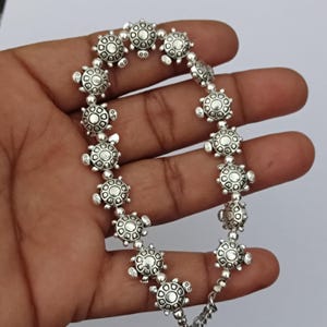 Könnte beinhalten: Ein silberfarbenes Armband mit einer Reihe sich wiederholender schildkrötenförmiger Anhänger. Jeder Schildkrötenanhänger weist ein detailliertes Panzermuster mit einem zentralen kreisförmigen Element und kleinen Beinen auf, die in einer Kette verbunden sind.