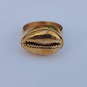 Peut inclure: Une bague dorée ornée d'un coquillage cauri en son centre. Le coquillage présente une finition métallique polie avec une ouverture dentelée au milieu. L'anneau de la bague est lisse et circulaire, assorti à la teinte dorée du coquillage.