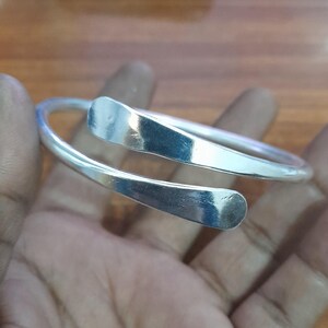May include: A silver-colored cuff bracelet with a curved, open design. The bracelet features two flat, rectangular ends that overlap slightly. The metal has a polished, reflective surface, and the bracelet appears to be made of a solid, durable material.