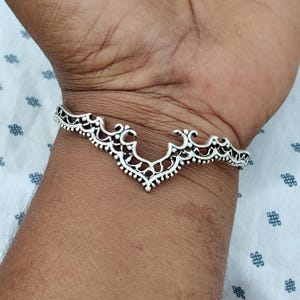 May include: A silver bracelet with an openwork design. The bracelet has an intricate pattern with a V-shaped central element and decorative edges. The bracelet is worn on a wrist.