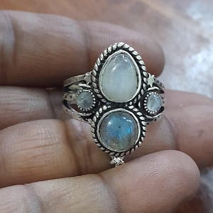 May include: A silver ring with a central pear-shaped moonstone, a round labradorite, and two smaller moonstones. The ring has a detailed, textured design with a rope-like border and star accents, showcasing a vintage or bohemian style.