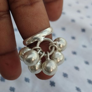 May include: A silver ring featuring a cluster of seven small, round bells. The bells are suspended from a central ring attached to the main band of the ring. The ring is made of a shiny, reflective metal.