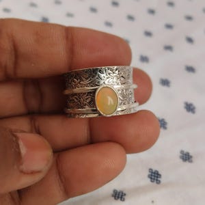 Puede incluir: Un anillo de plata con una piedra preciosa de ópalo ovalada. El anillo tiene un patrón floral y una banda giratoria. El ópalo es de color amarillo pálido. El anillo se sostiene en una mano.
