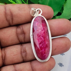 May include: An oval-shaped pendant featuring a faceted, deep pinkish-red gemstone set in a polished silver-toned metal frame. The pendant includes a simple silver-toned bail at the top for attachment to a chain or cord, showcasing a smooth finish.