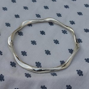May include: A silver bangle with a flowing, irregular shape. The bracelet is circular and made of a shiny metal. The background is a white fabric with a repeating pattern of small, dark blue floral shapes.